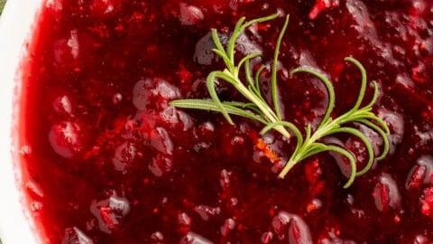 Close-up of homemade cranberry sauce in a beautiful white ceramic bowl, garnished with candied orange peel and fresh rosemary sprigs on a rustic wooden table