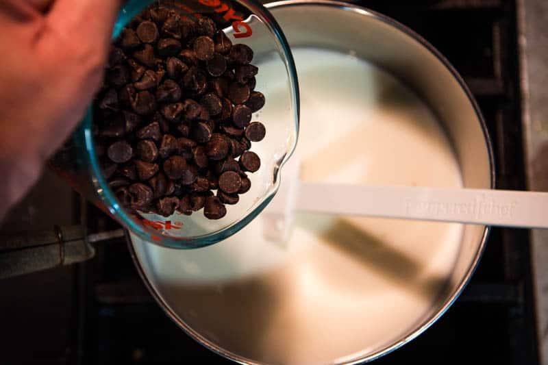 Hands stirring rich dark chocolate chips into warm milk in a saucepan on a stove