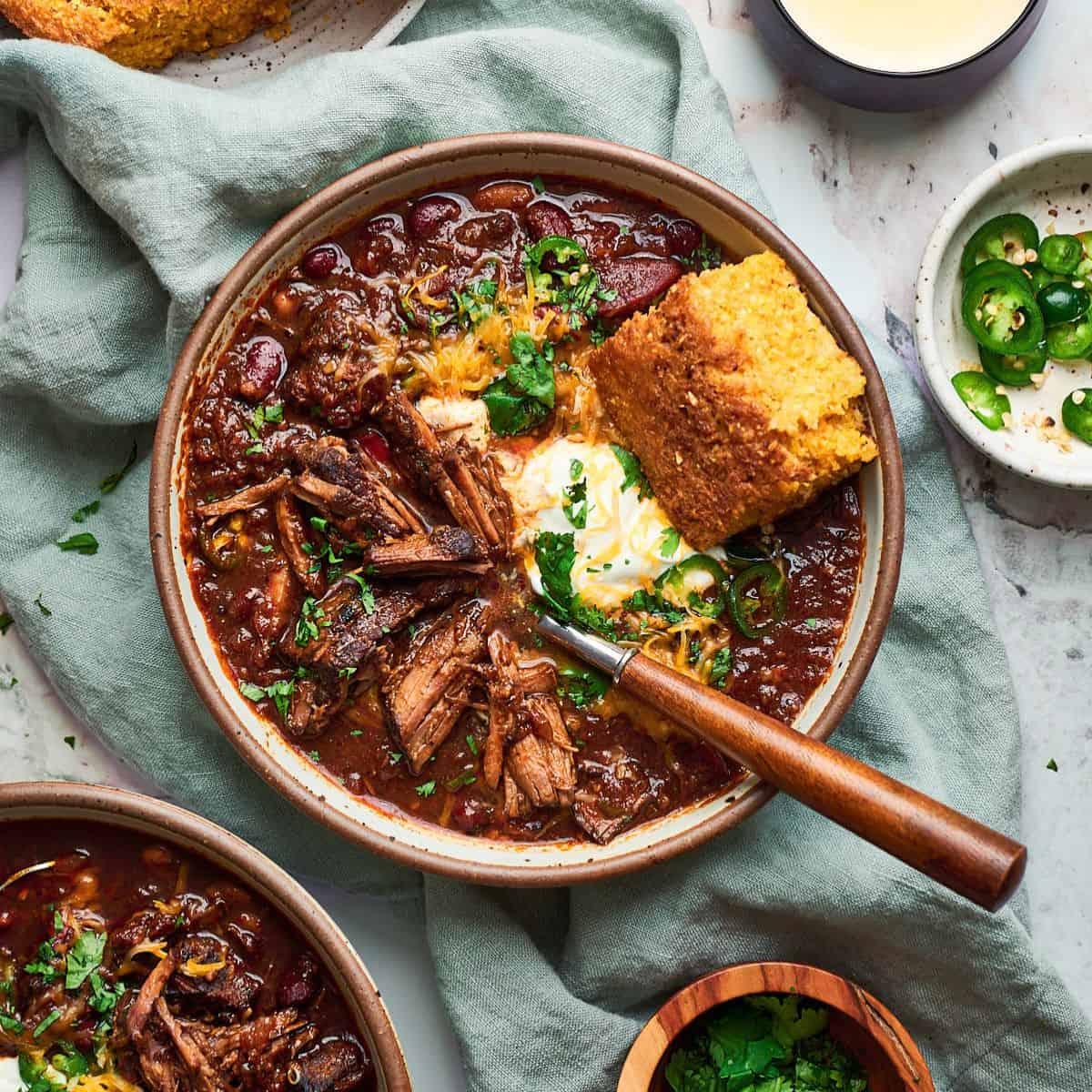 Ingredients for chili beef roast laid out on a rustic wooden cutting board: chuck roast, chili powder, cumin, diced tomatoes, chipotle peppers, cocoa powder, fresh garlic, onion