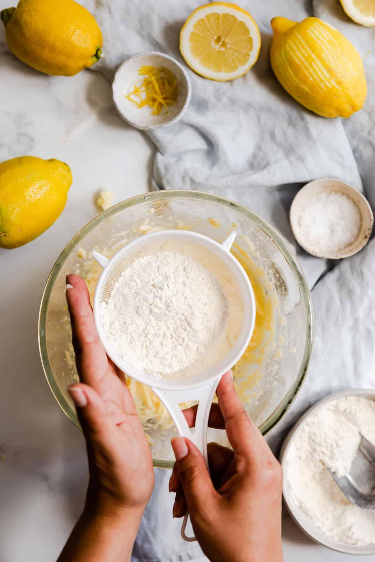 overhead shot of a baker zesting a fresh lemon over a bowl of cake batter, with other baking ingredients visible on a light kitchen counter