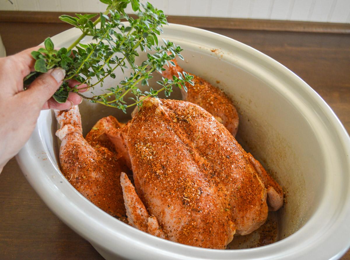 hands placing ingredients into a large slow cooker, colorful spices and chicken visible
