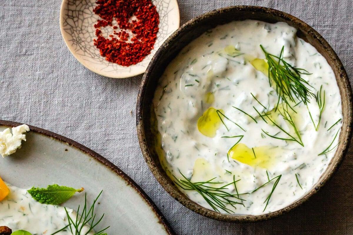 Assortment of fresh herbs like dill, mint, and parsley on a wooden cutting board with a bowl of Greek yogurt