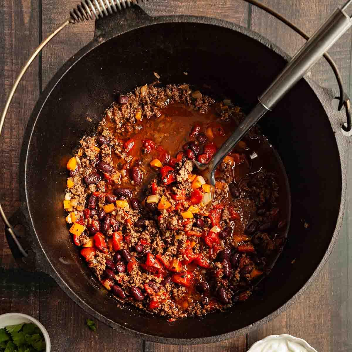 A person stirring chili-fired ground beef in a large cast iron skillet, steam rising, vibrant spices visible.