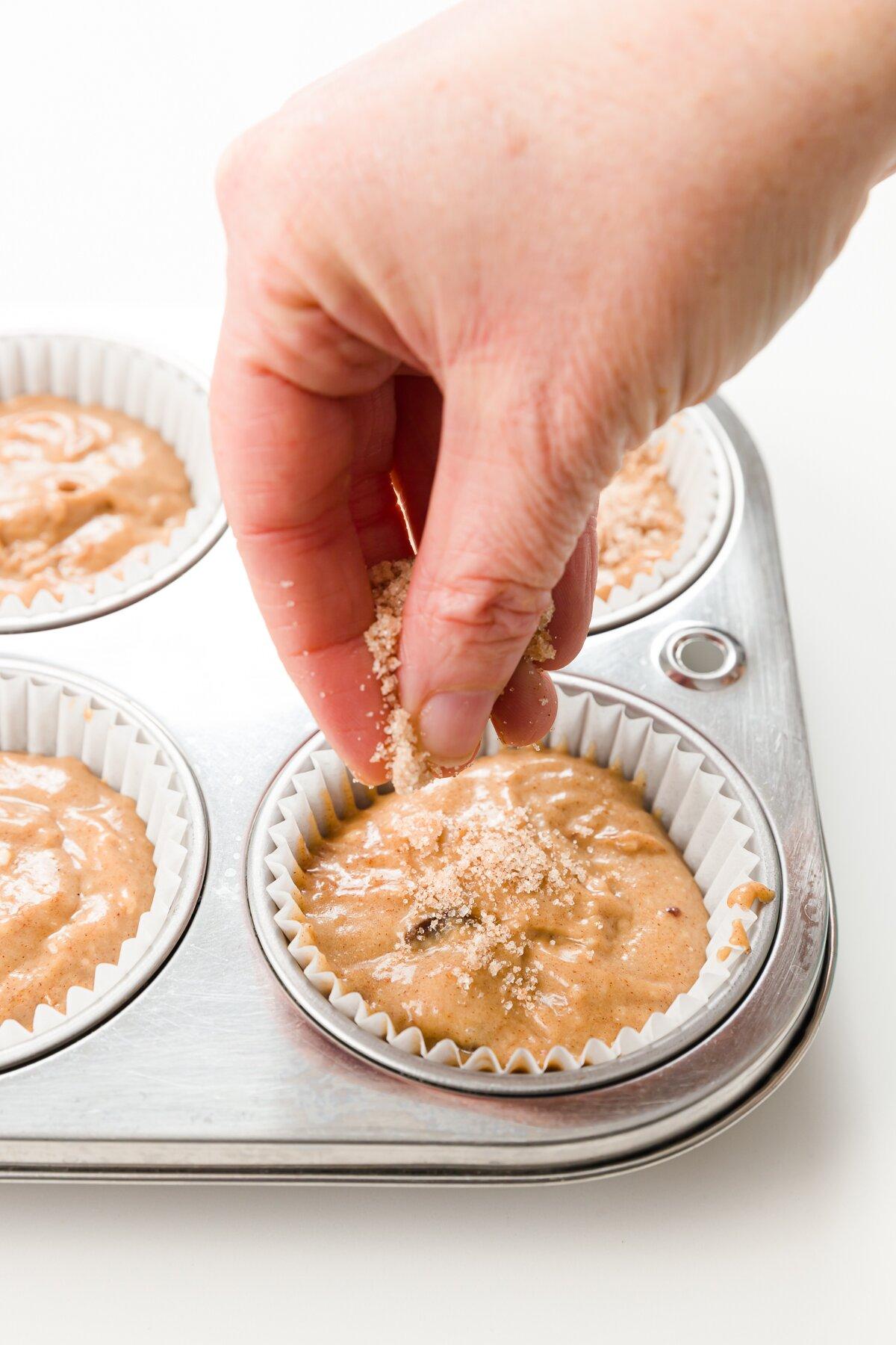 a close-up of a hand sprinkling cinnamon on muffin batter in a muffin tin