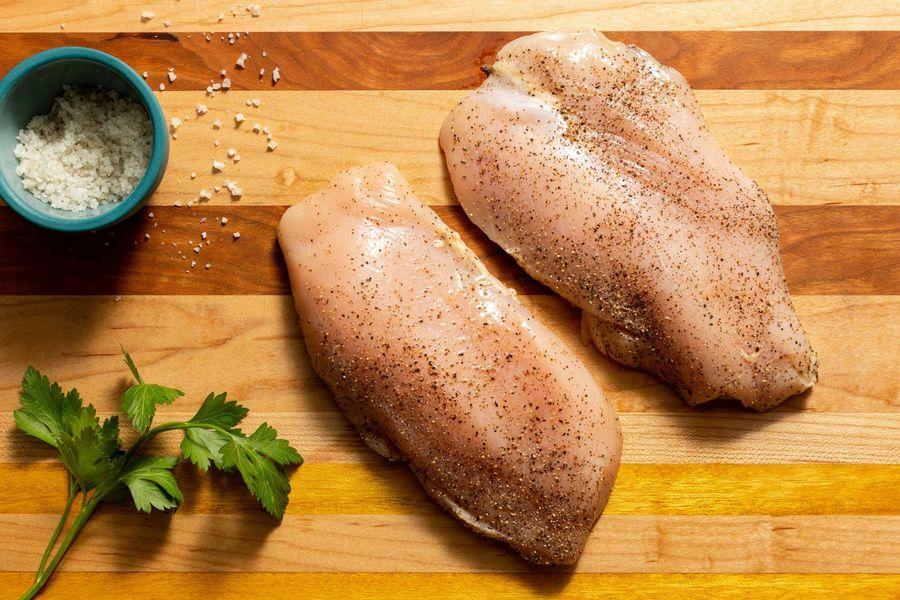 Fresh chicken breasts being seasoned with salt and pepper on a cutting board