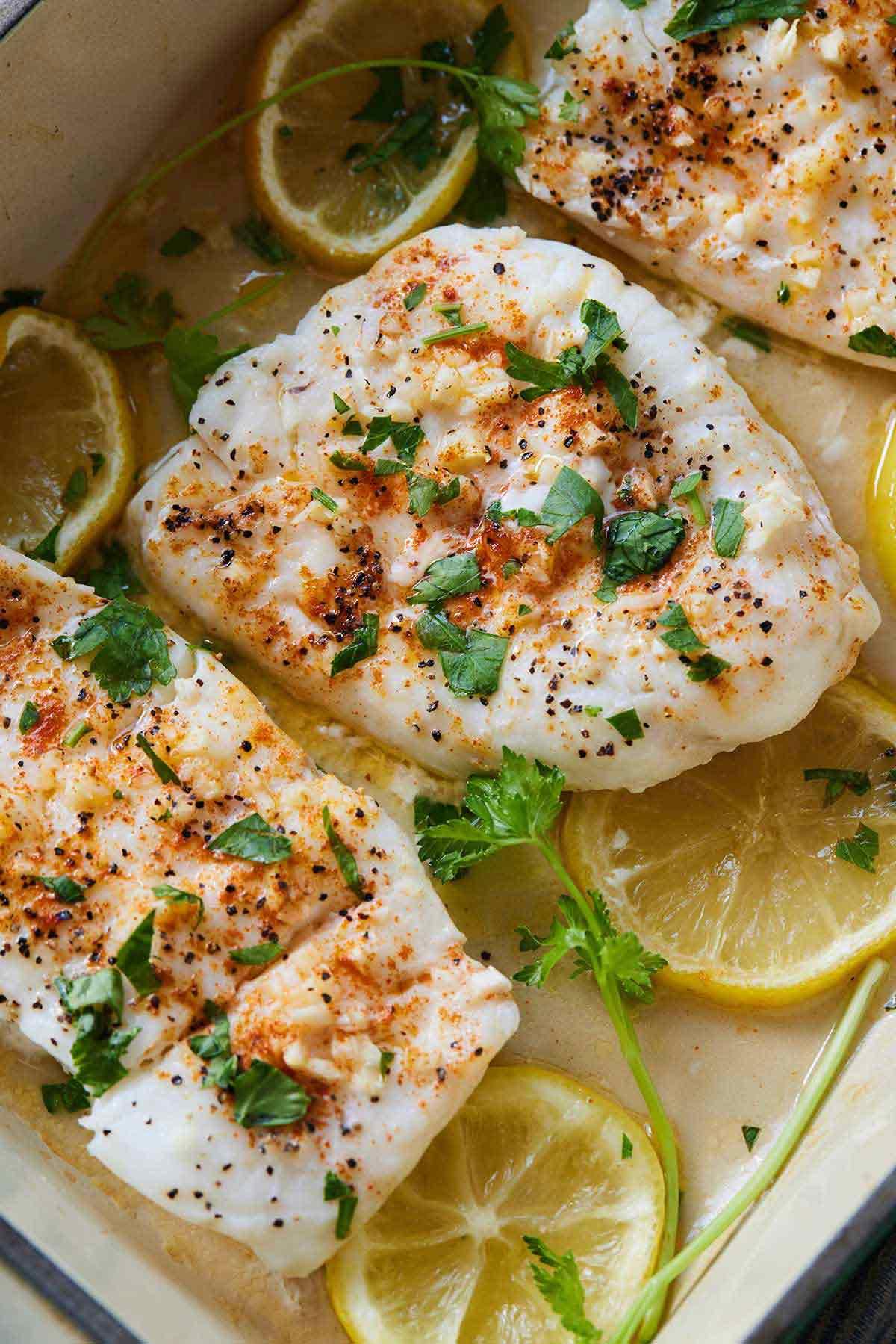 Baked cod with lemon slices and herbs in a white baking dish, fresh and inviting, golden crust, steam rising