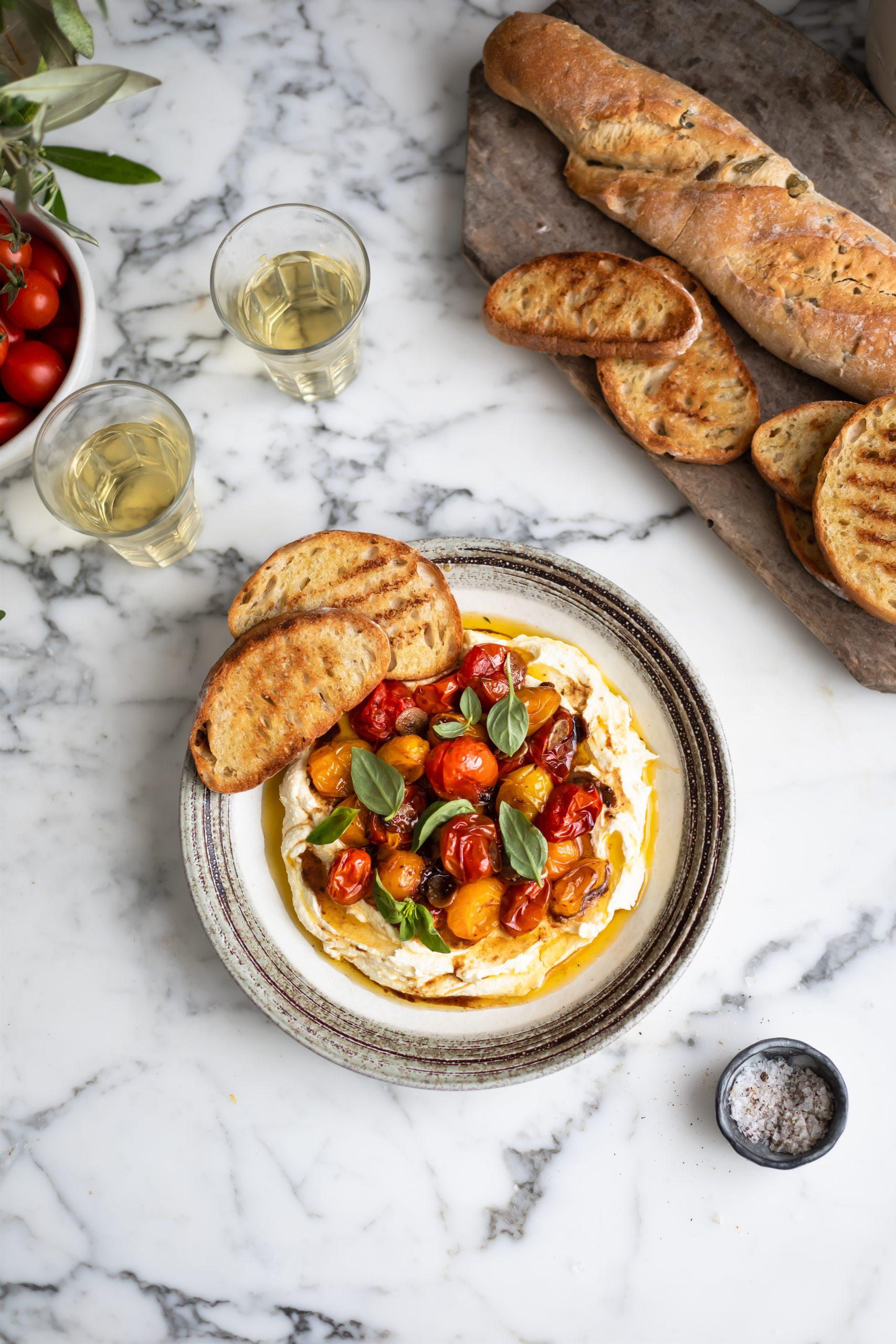 Close-up of creamy whipped feta dip garnished with roasted cherry tomatoes, fresh basil leaves, and a drizzle of olive oil, served with toasted pita bread and cucumber slices