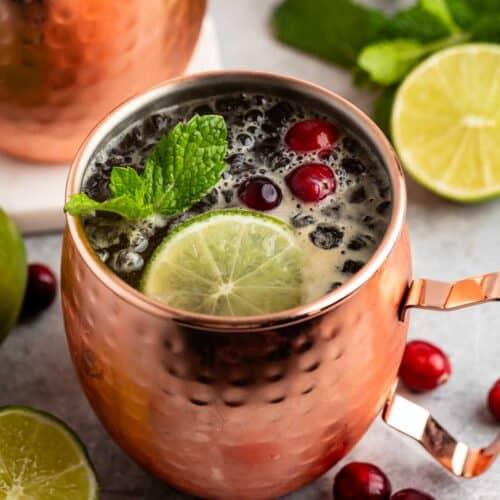 hands mixing a cranberry mule in a shiny copper mug, garnished with frosty lime slices and fresh cranberries, set against a blurred festive background