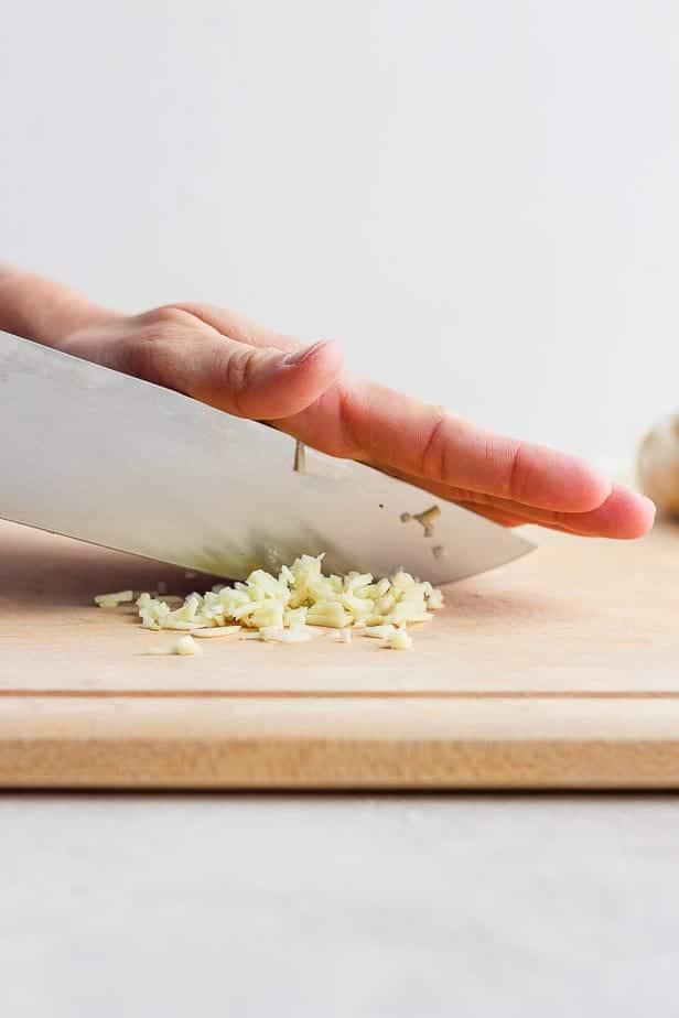 garlic cloves being minced on a cutting board next to a block of parmesan cheese