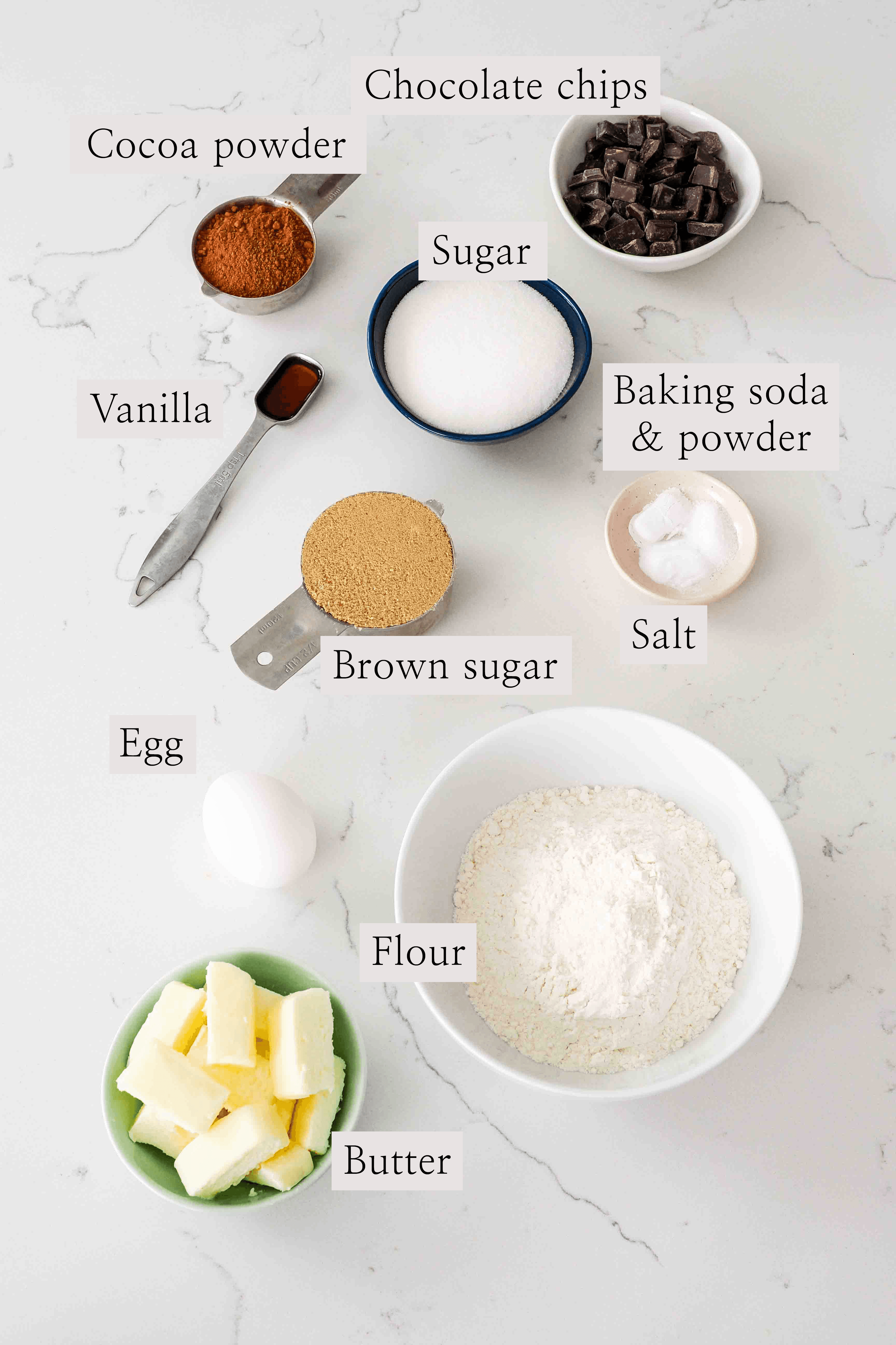 Ingredients for double chocolate cookies laid out on a marble counter, including cocoa powder, chocolate chips, butter, and brown sugar.