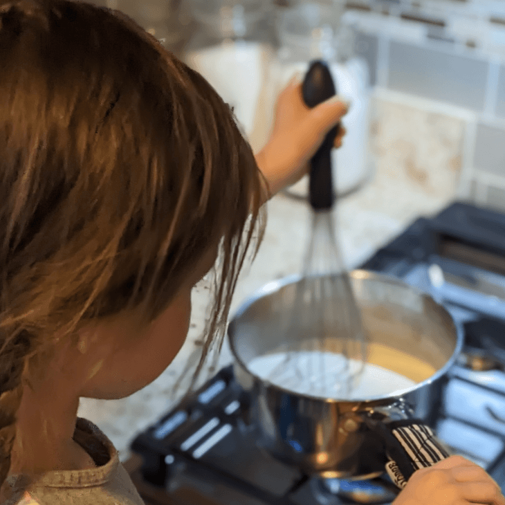Hands stirring a creamy white sauce in a saucepan on a stove, with specks of black pepper visible