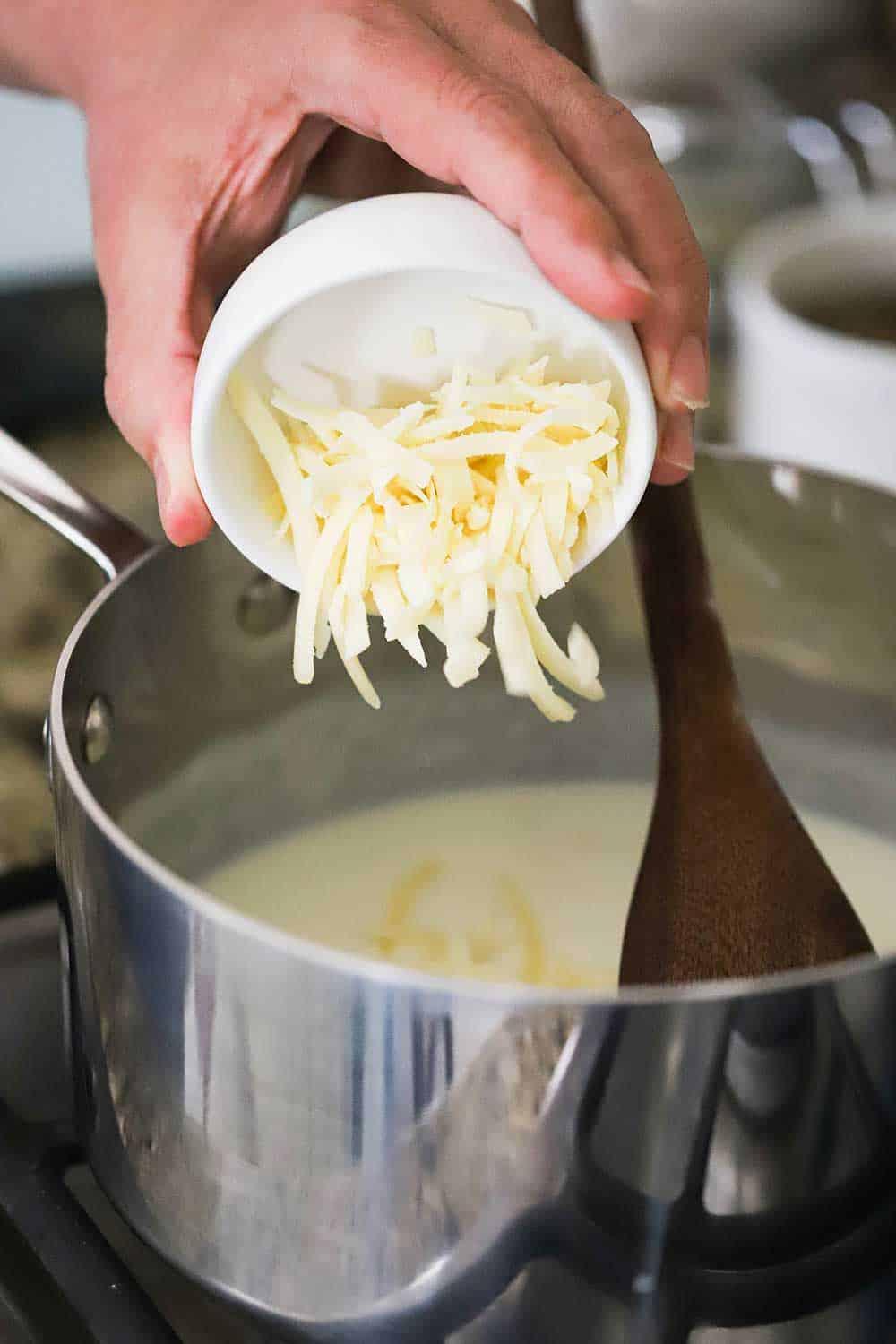 Hands grating Gruyere cheese into a large pot of bubbling béchamel sauce on a stovetop