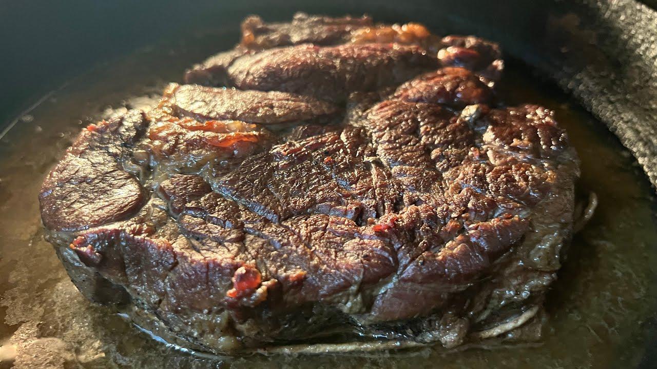 Close-up of a chuck roast being seared in a hot cast-iron skillet, developing a rich brown crust, with visible steam and sizzle