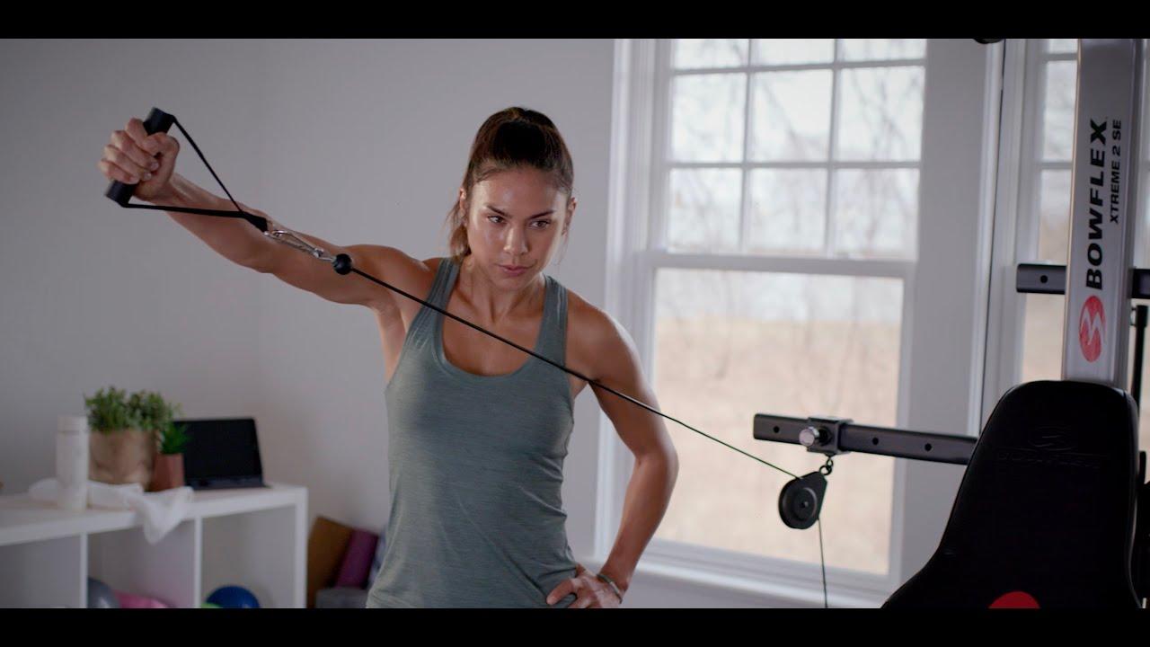 A woman smiling and looking fit, standing in front of the Bowflex Xtreme 2 SE Home Gym