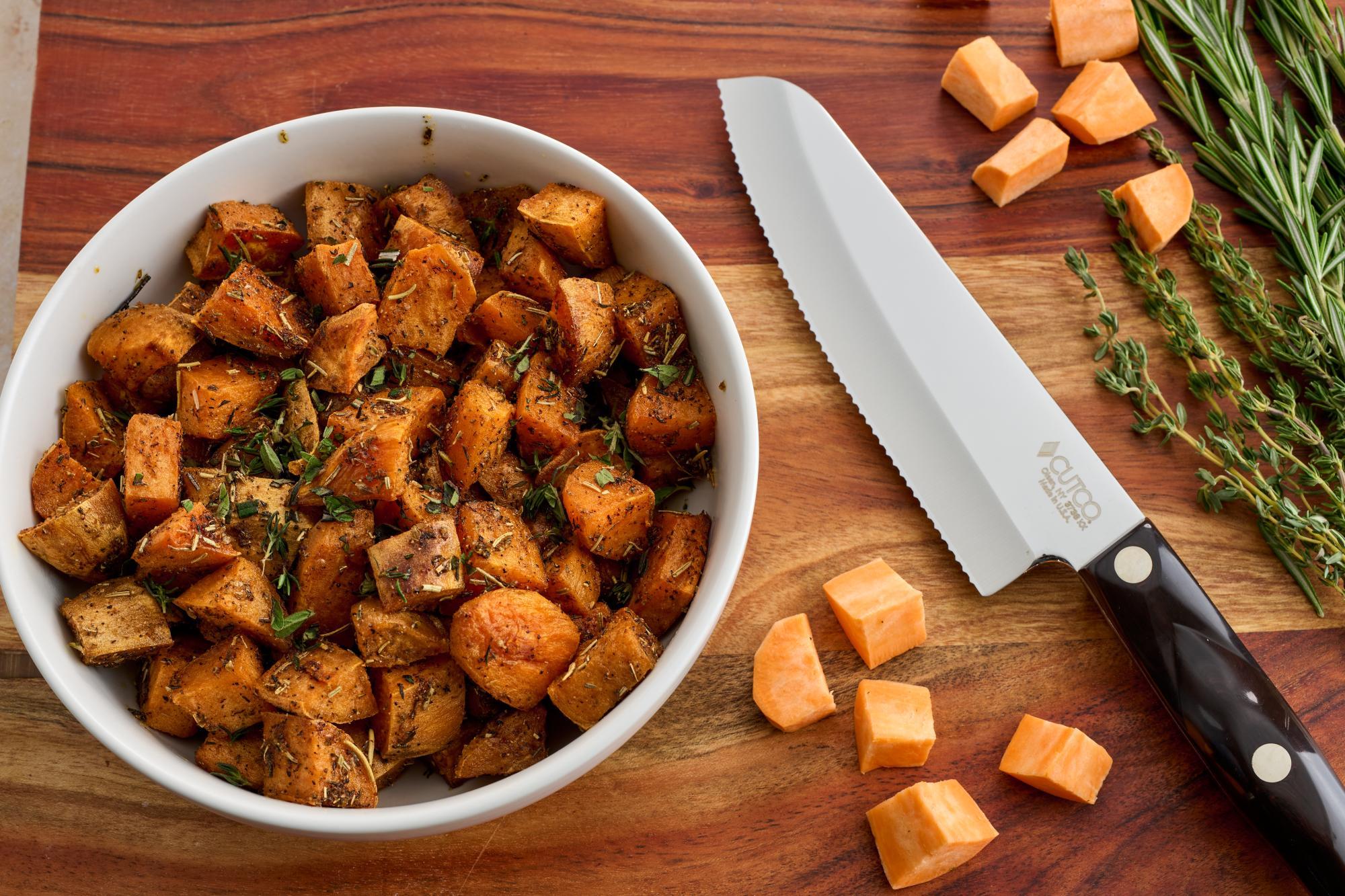 Fresh rosemary and thyme sprigs, garlic cloves, and cubed sweet potatoes on a wooden cutting board, pre-preparation shot, vibrant colors
