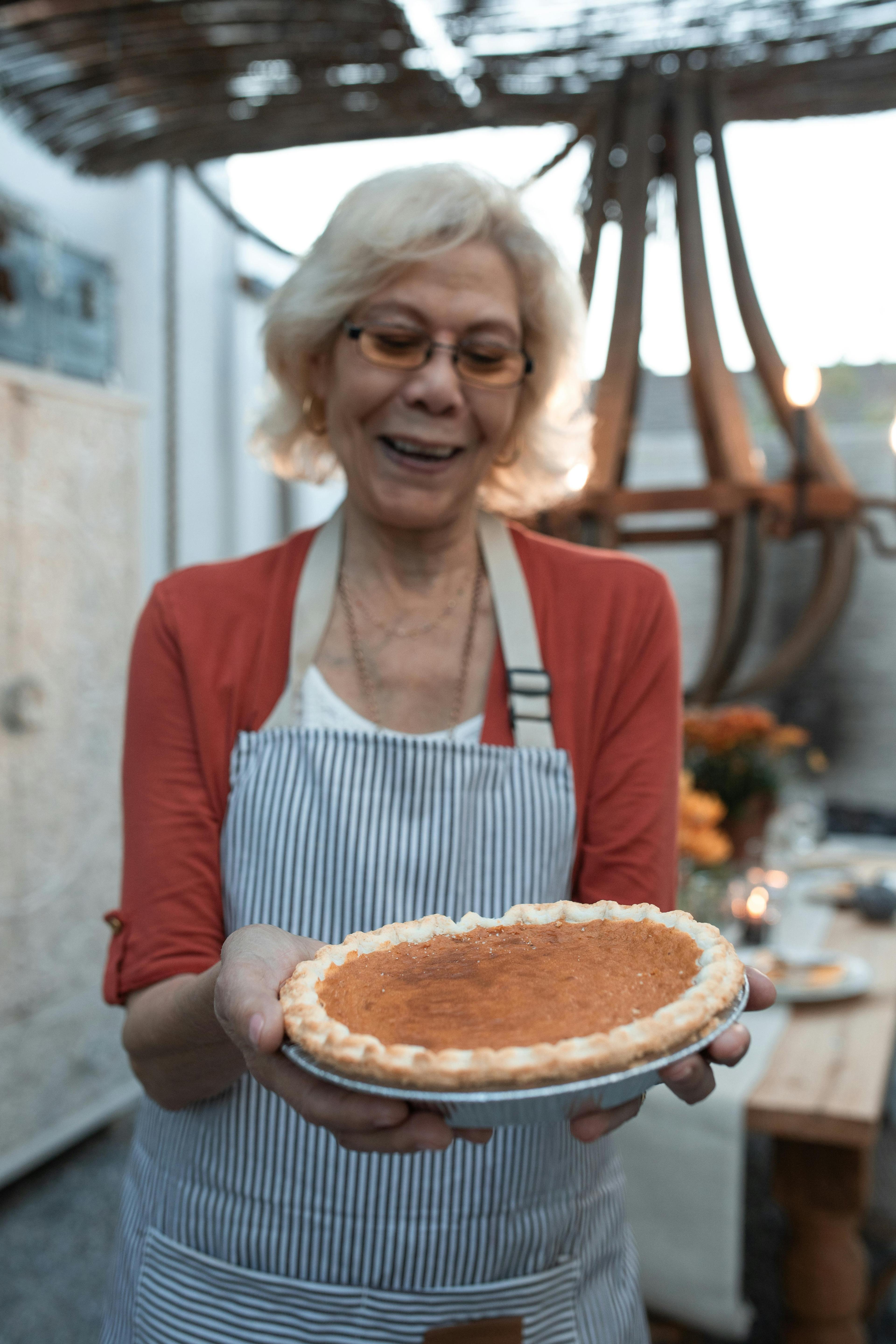Smiling woman in apron holding a freshly baked pumpkin pie, steam gently rising, in a cozy kitchen setting