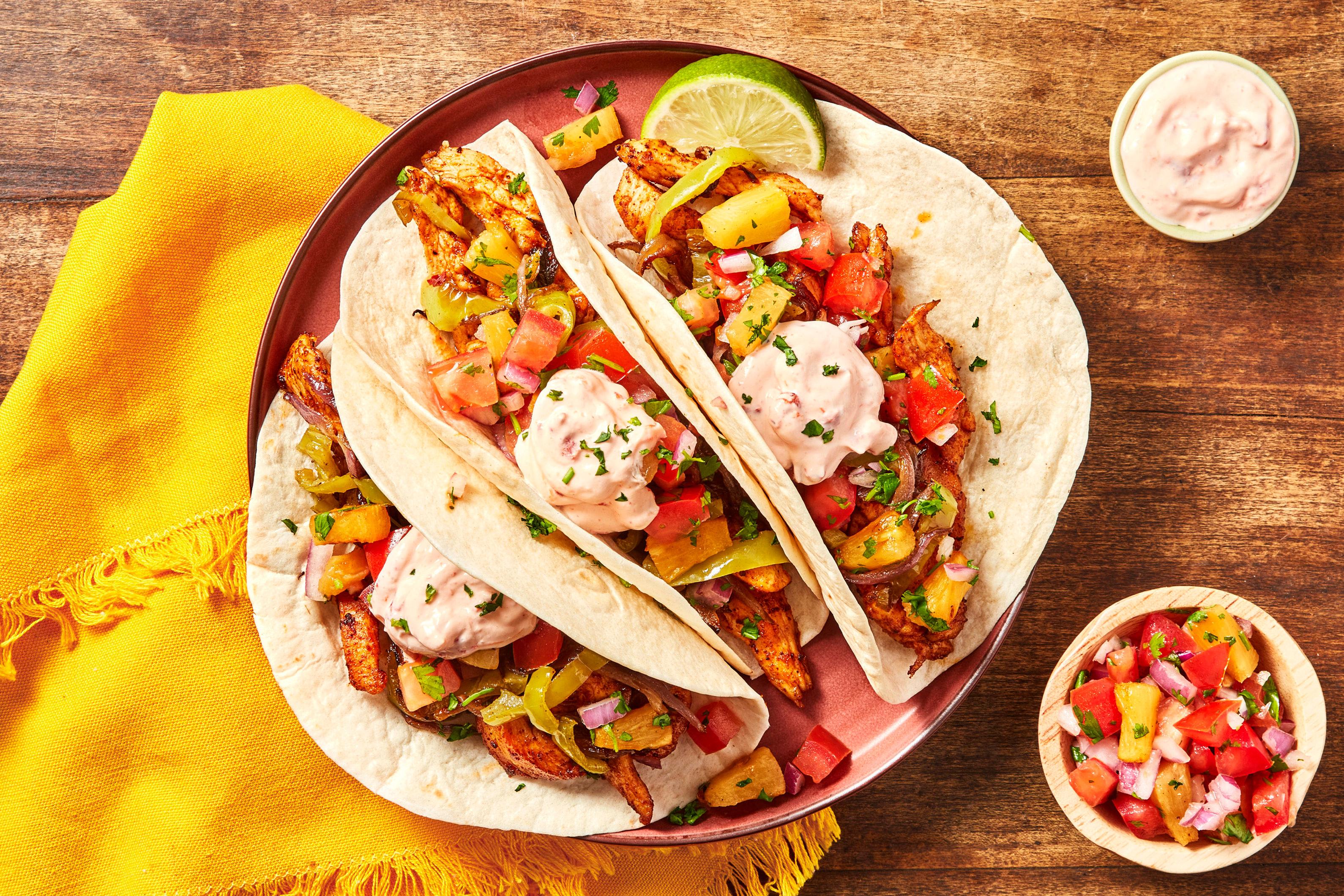 Overhead shot of assembled pineapple chicken tacos on various colorful plates, with a blurred background of a happy family eating at a table