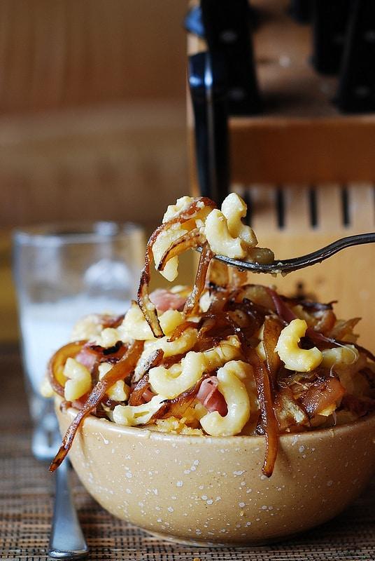 Person serving a generous portion of mac and cheese with caramelized onions and bacon
