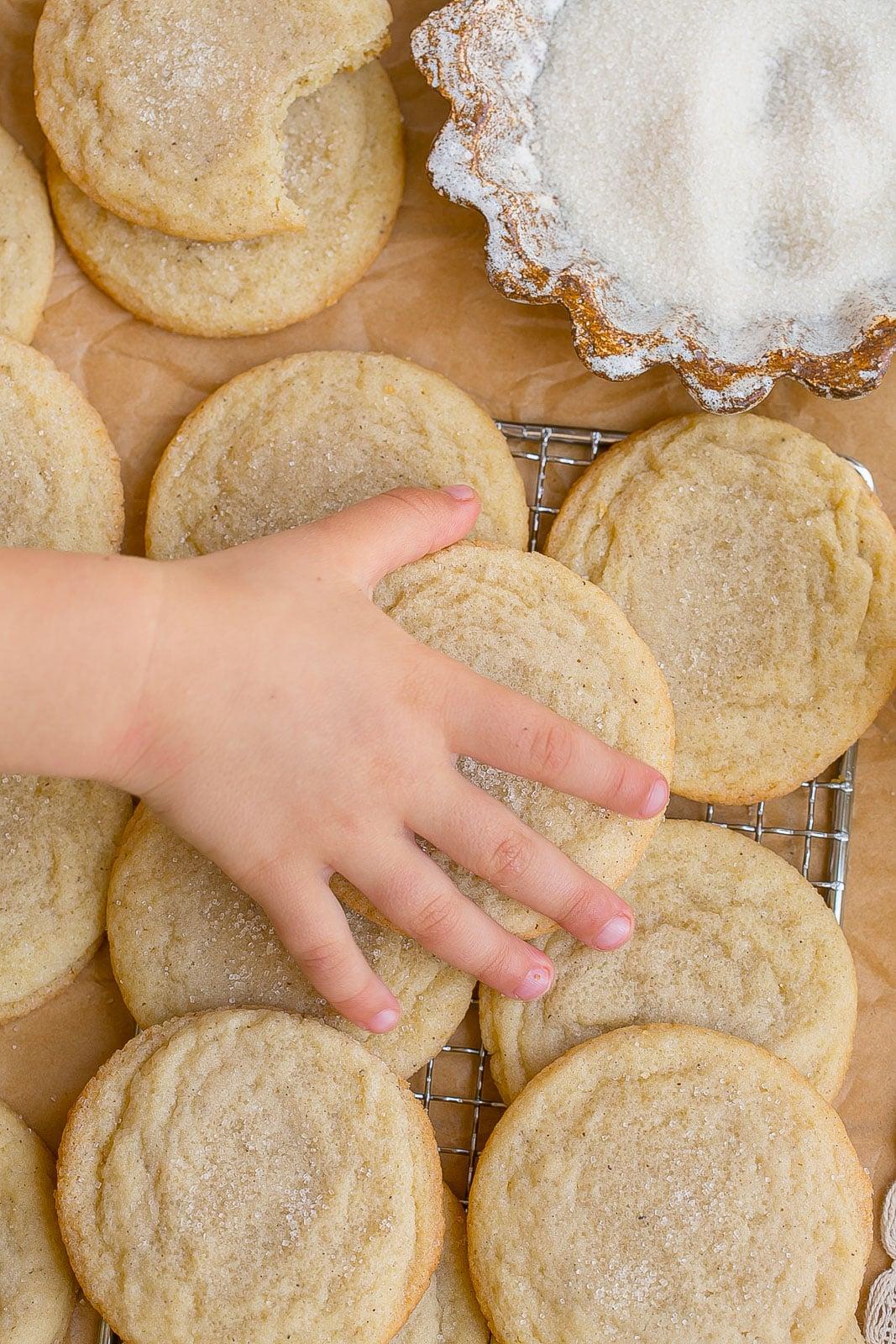 A child's hands reaching for vanilla bean speckled sugar cookies on a vintage baking tray, soft focus