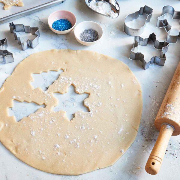 Hands rolling out sugar cookie dough on a floured counter with various Christmas cookie cutters nearby