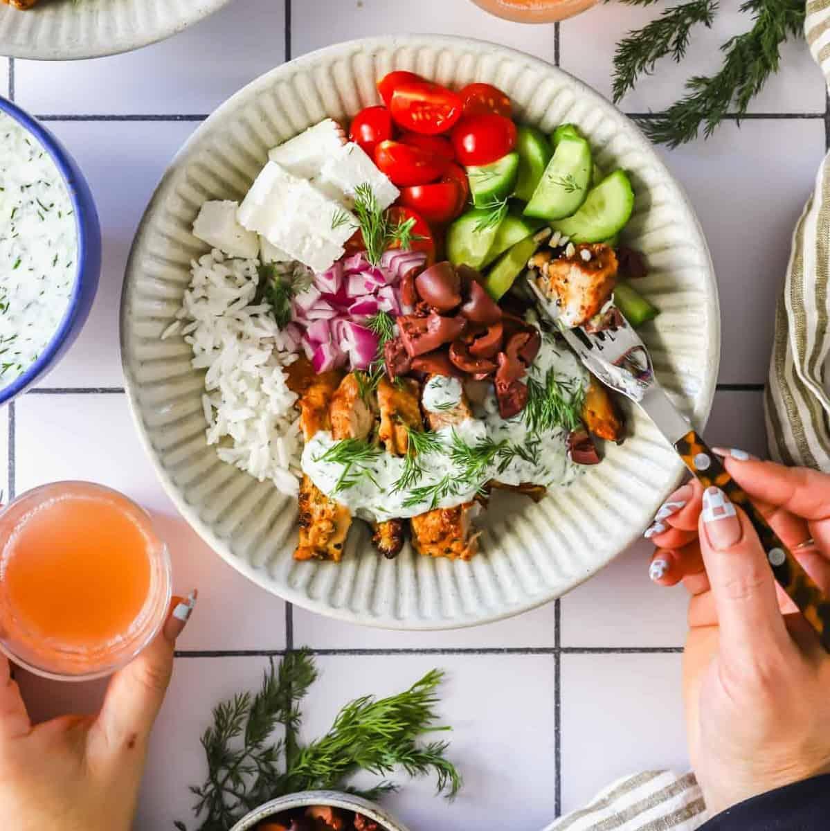 Hands assembling a colorful Greek chicken bowl with a focus on fresh vegetables and marinated chicken