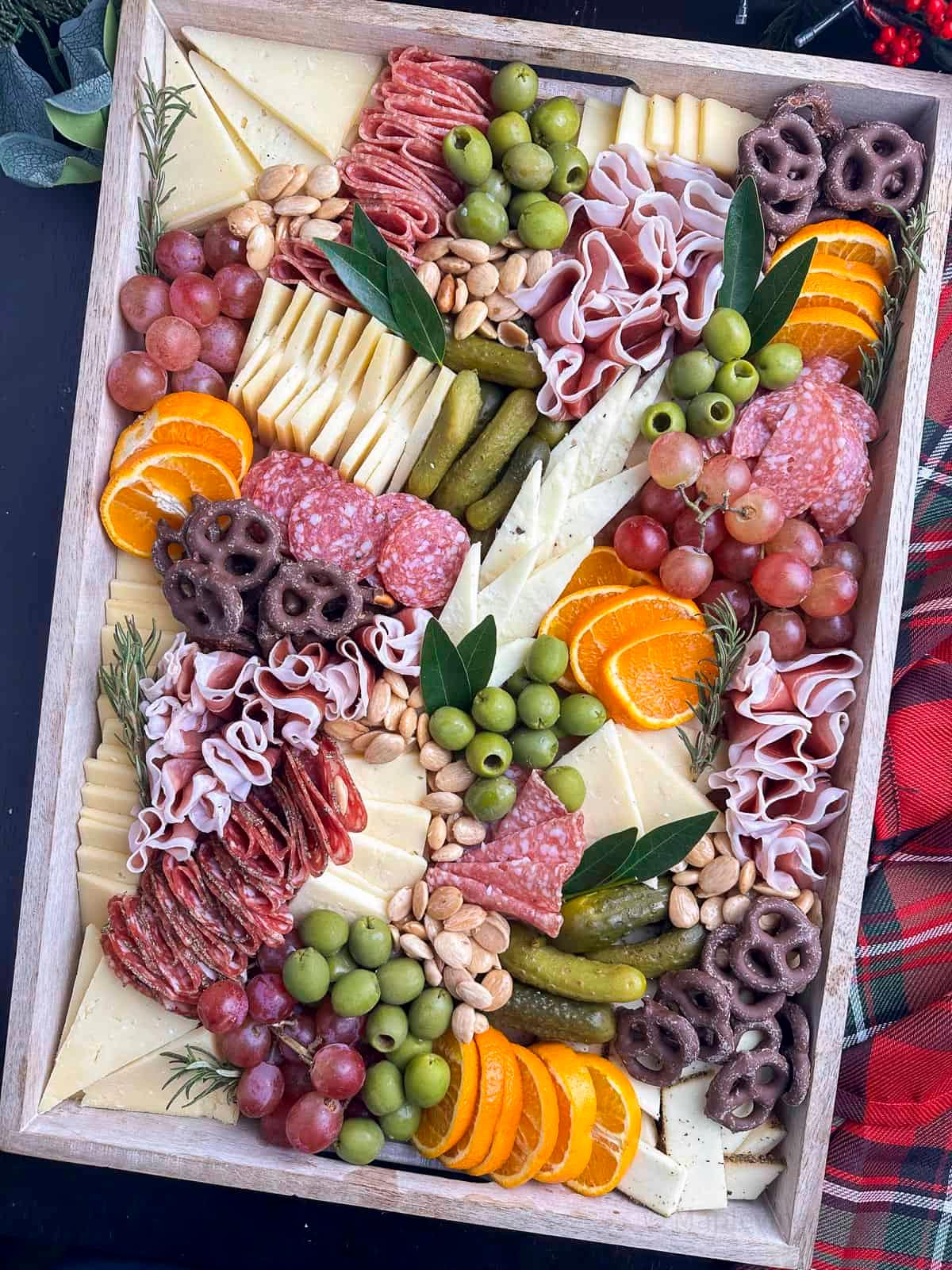 Hands arranging various elements on a large charcuterie board, including prosciutto, fig spread, and different cheeses