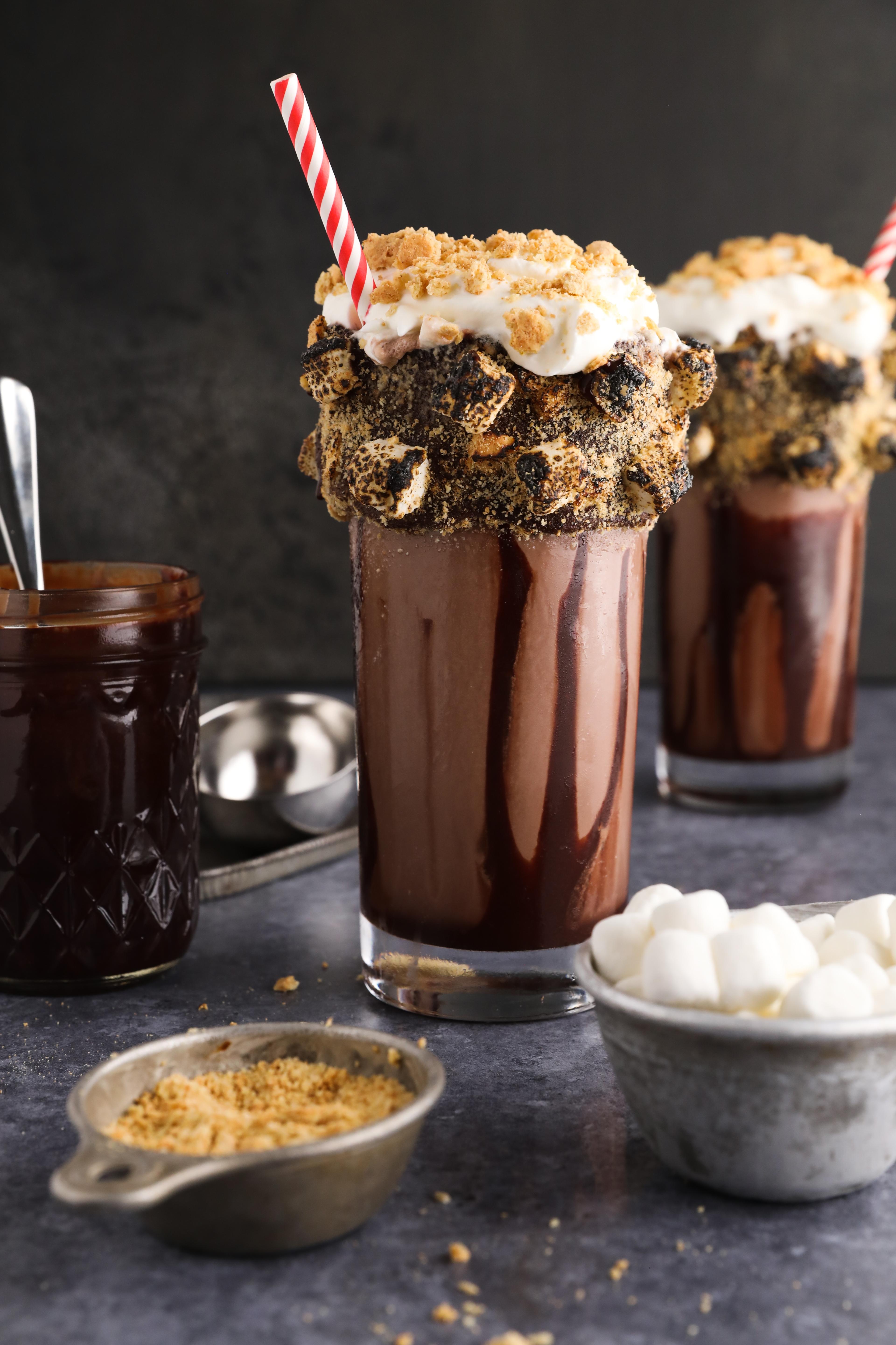 a beautifully layered milkshake with whipped cream, chocolate chunks, marshmallows, and raspberries cascading down the sides of the glass