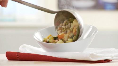 a person ladling minestrone soup into a bowl