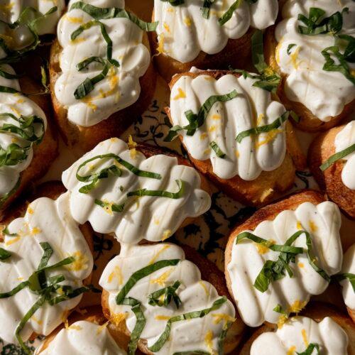 close-up of a hand reaching for a piece of golden crispy crostini topped with white whipped ricotta and a basil leaf