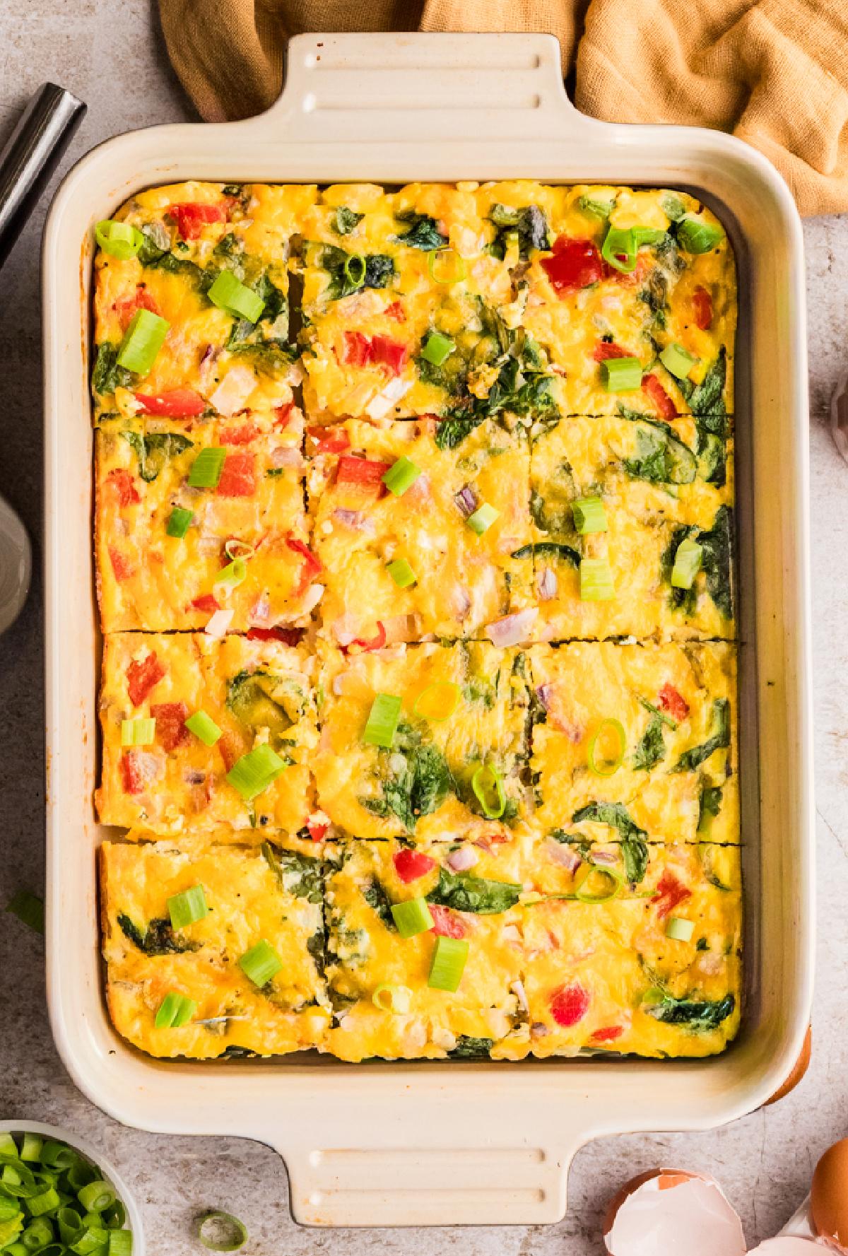 various fresh ingredients for an egg bake laid out on a kitchen counter: eggs, shredded cheese, colorful bell peppers, fresh spinach, a carton of milk