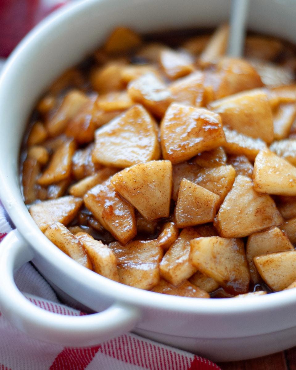 Hands tossing sliced apples with cinnamon and sugar in a large bowl, cozy kitchen setting.