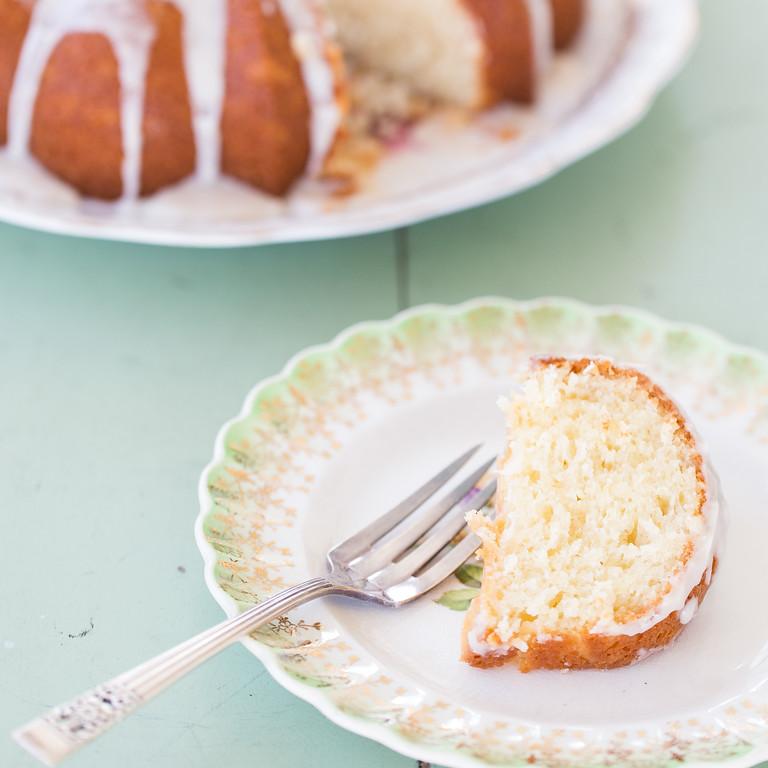 close-up of a slice of light lemon yogurt Bundt cake on a white plate with a fork, showing its delicate crumb and a shiny lemon glaze, garnished with a curl of lemon zest and a small sprig of mint