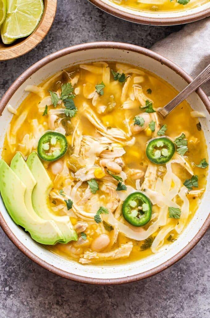 variety of chili toppings in small bowls: cilantro, sour cream, avocado, and tortilla chips next to a serving of white chicken chili