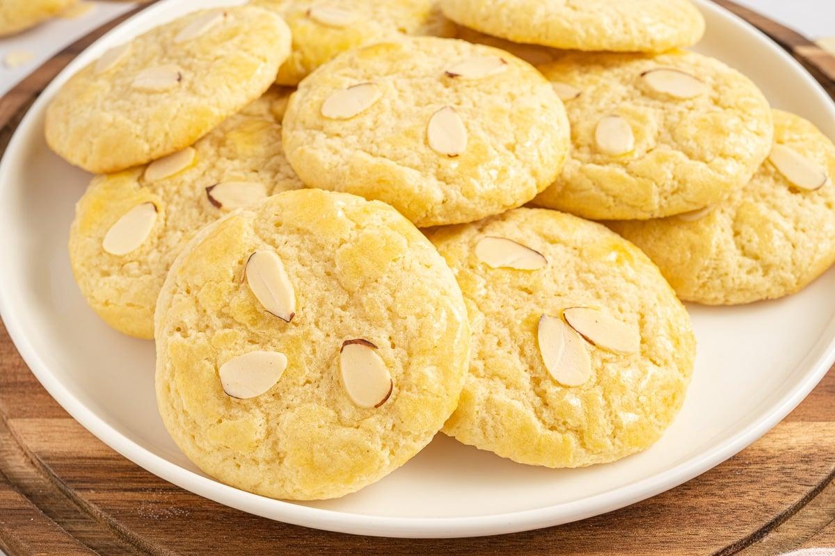 person laughing happily while holding a plate of perfectly baked golden almond cookies with crisp edges, with a soft blur of a home kitchen in the background