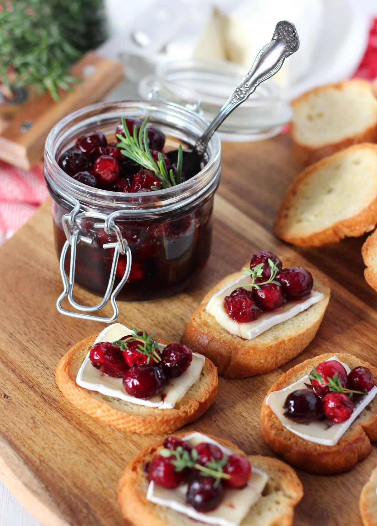 Small jar of homemade cranberry sauce on a rustic wooden board with a silver spoon, next to a piece of toasted brioche.