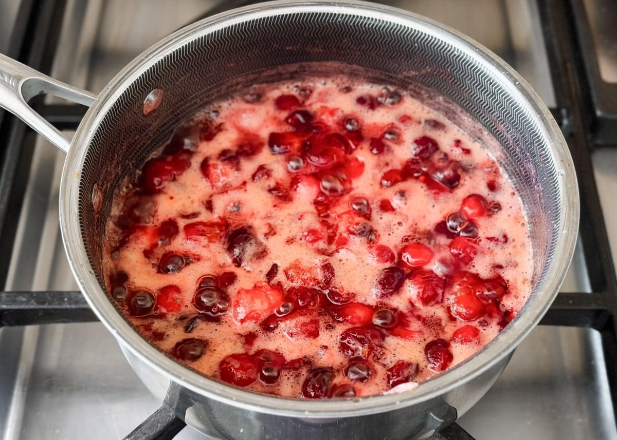 Cranberry sauce simmering gently in a saucepan on a stove, with steam rising and cranberries bursting