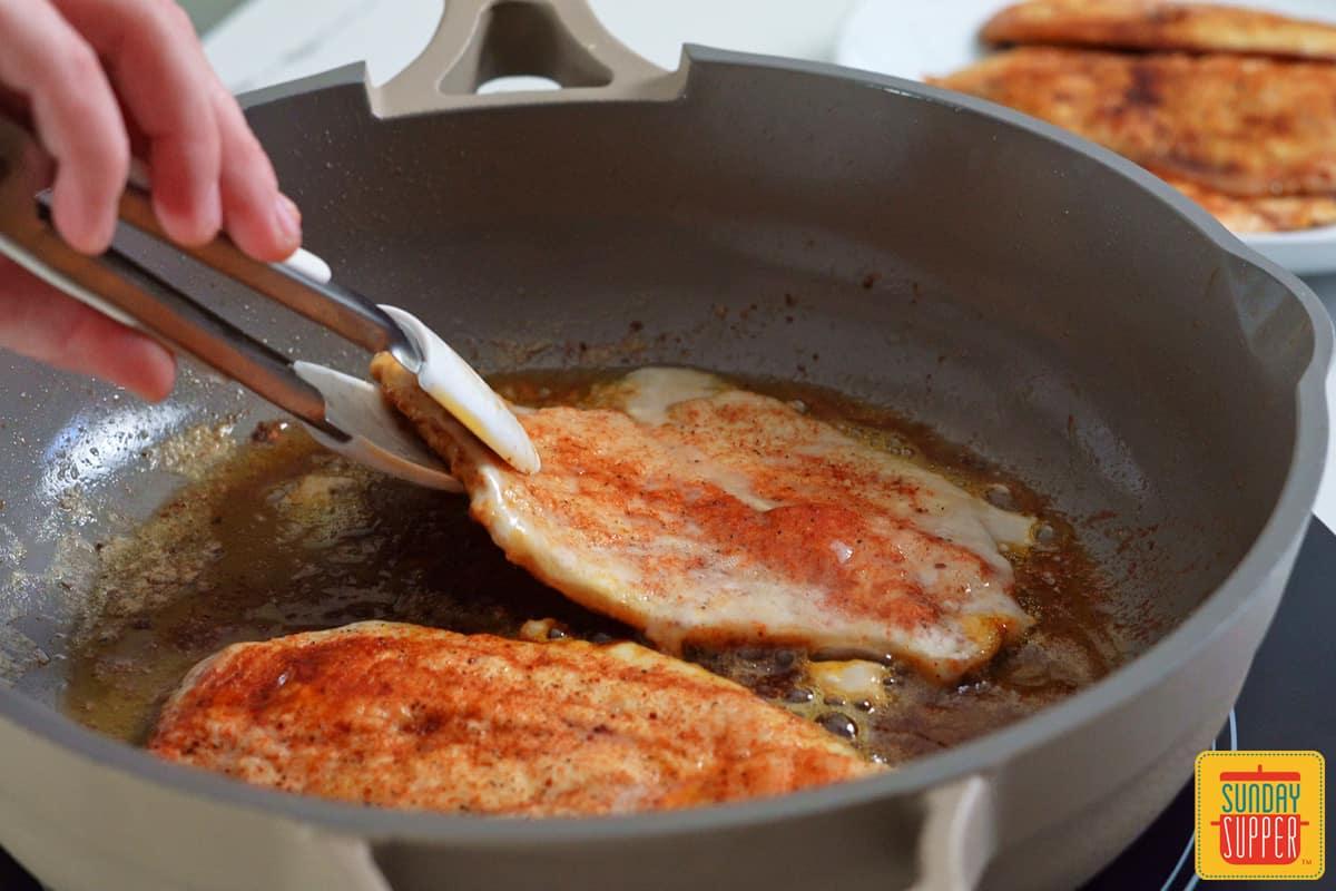 Hand holding a spatula, flipping a chicken breast in a hot skillet with melted butter