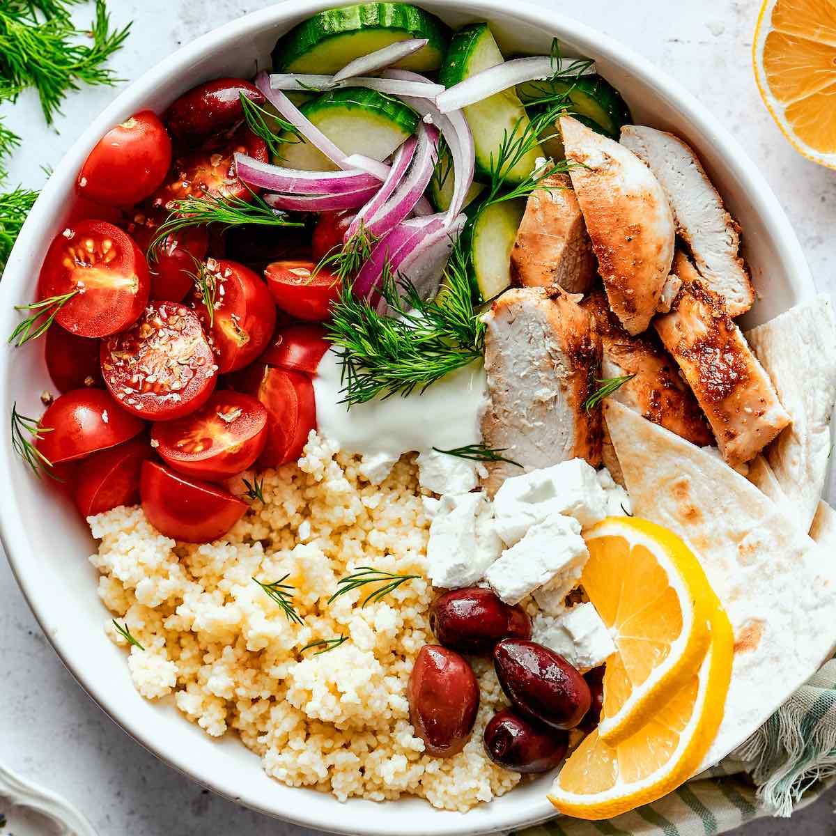 Smiling person enjoying a vibrant Greek chicken bowl on a sunlit patio, with a coastal background