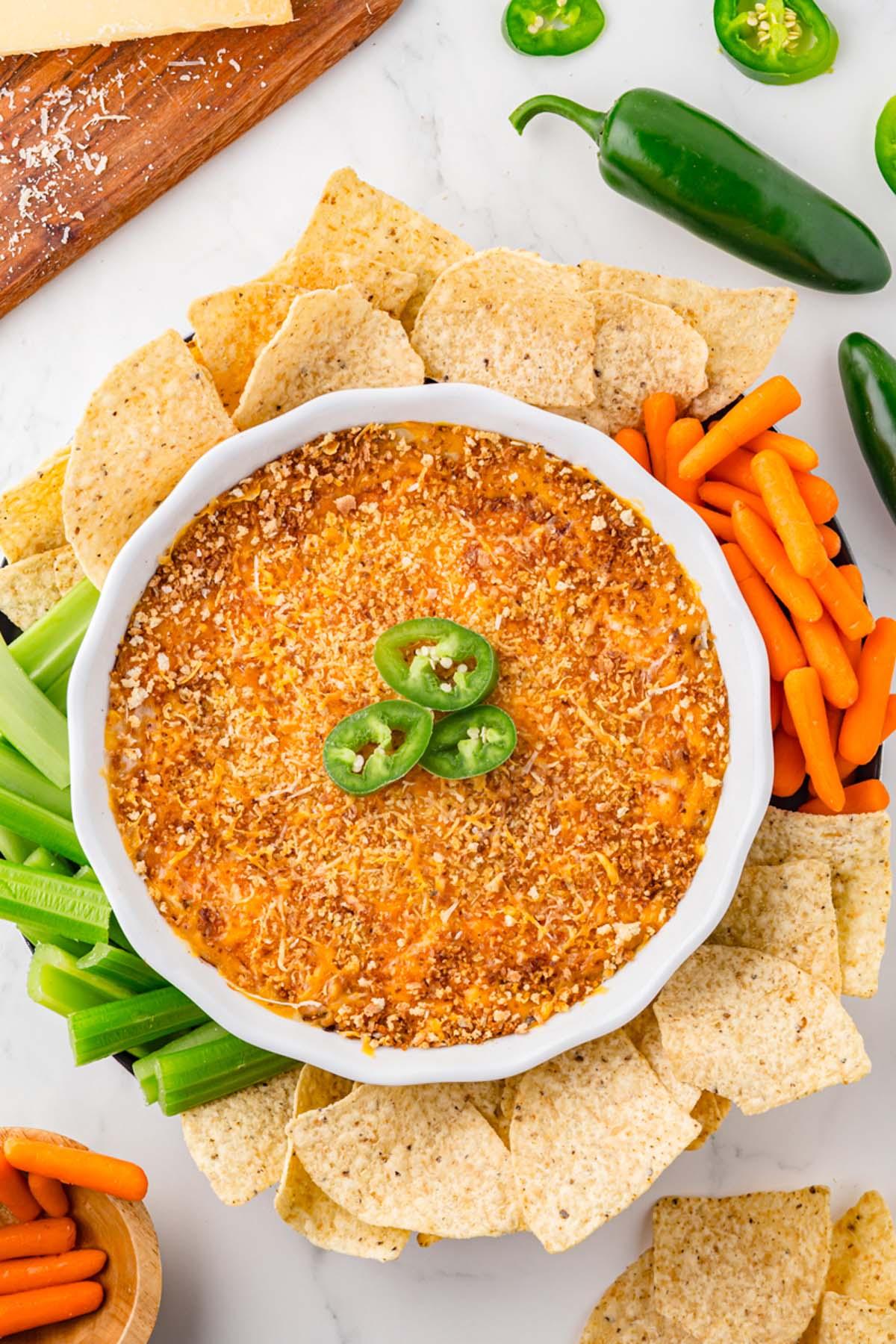 Overhead shot of a bubbling hot jalapeño popper dip in a ceramic dish, surrounded by golden-brown tortilla chips and fresh celery sticks.