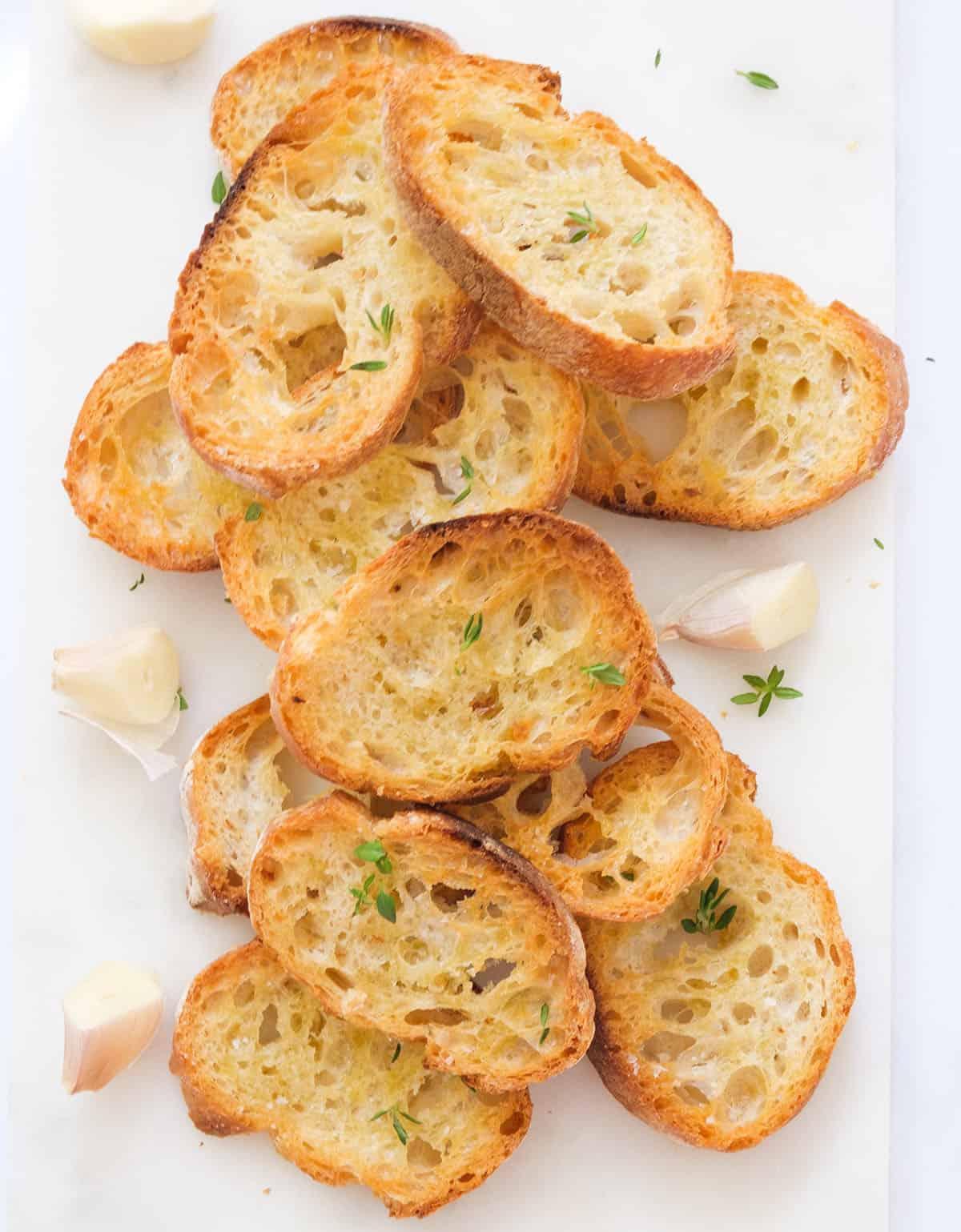 stack of golden-brown crispy crostini slices on a cutting board, with a garlic clove nearby
