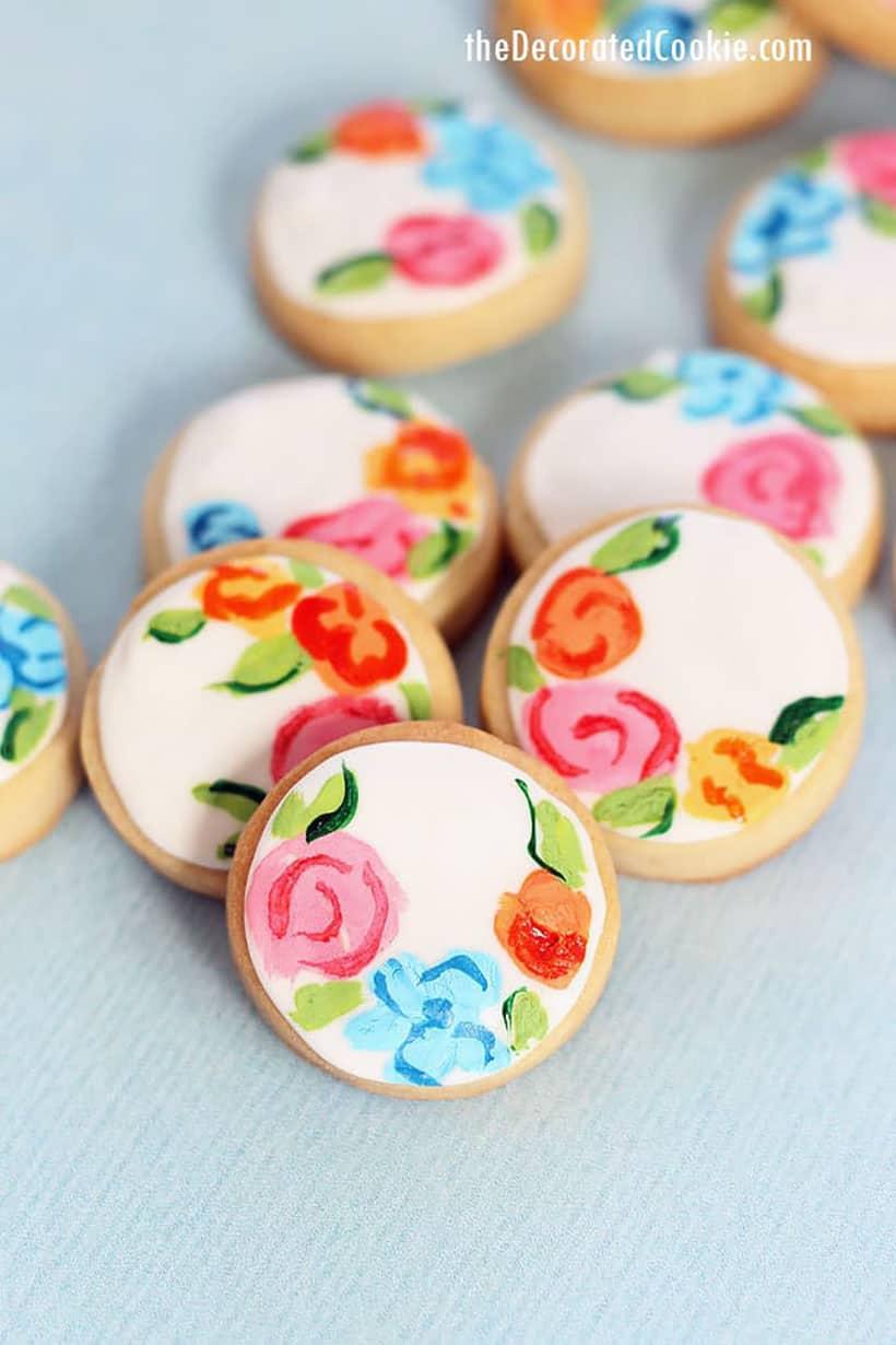 A person delicately painting a small flower design onto a round butter cookie with a fine-tipped brush