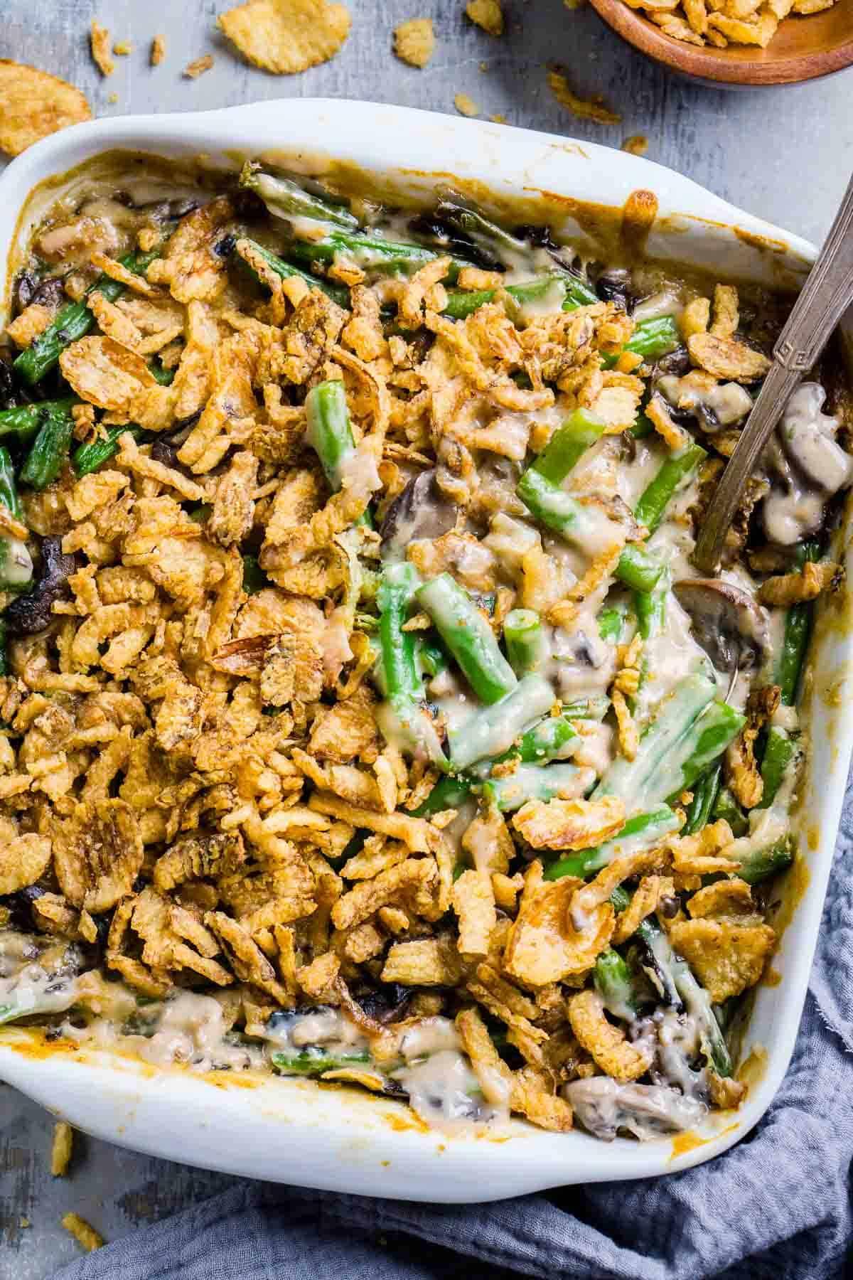 Homemade creamy green bean casserole with golden crispy onions on top, a Thanksgiving table setting in the background, warm inviting light