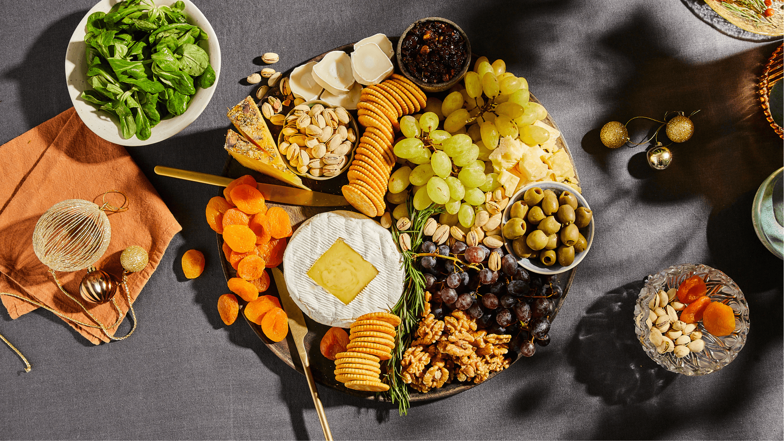 Overhead shot of a beautifully arranged charcuterie board centered around a baked camembert, with hands reaching for food