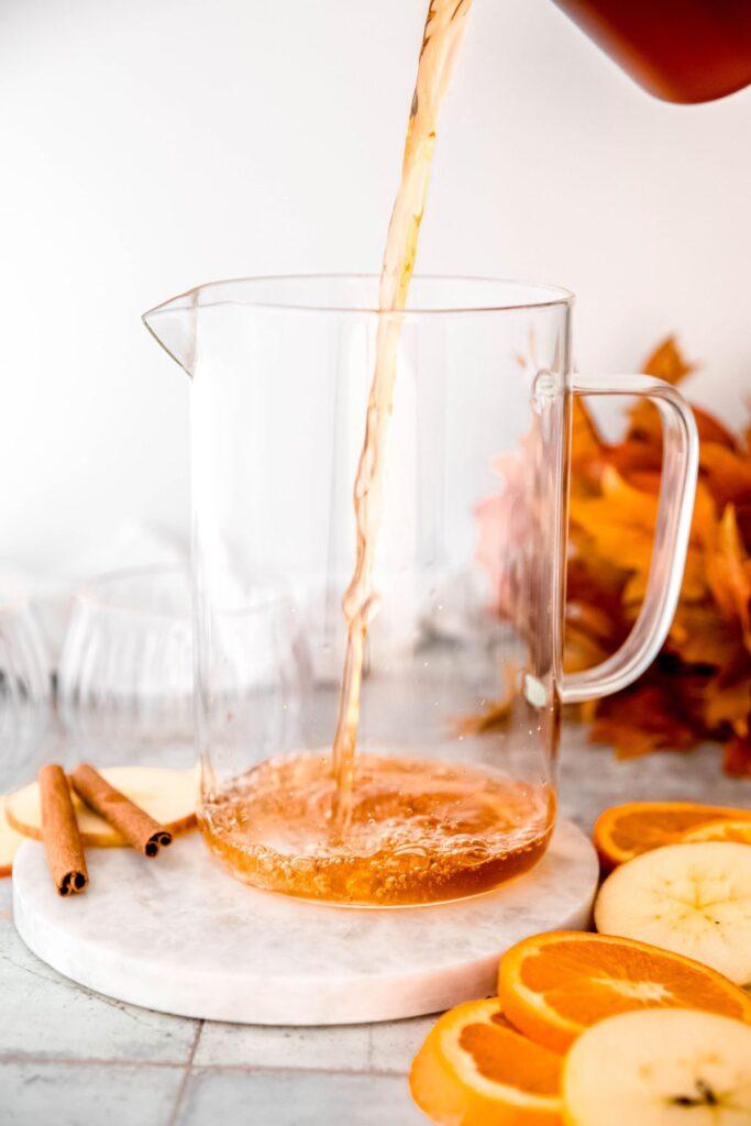 Hand pouring orange clove punch from a pitcher into a decorative glass mug