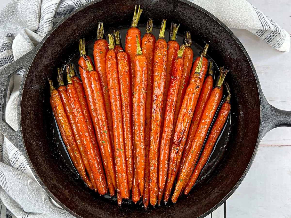 Hands gently stirring honey glazed carrots in a cast iron skillet, steam rising
