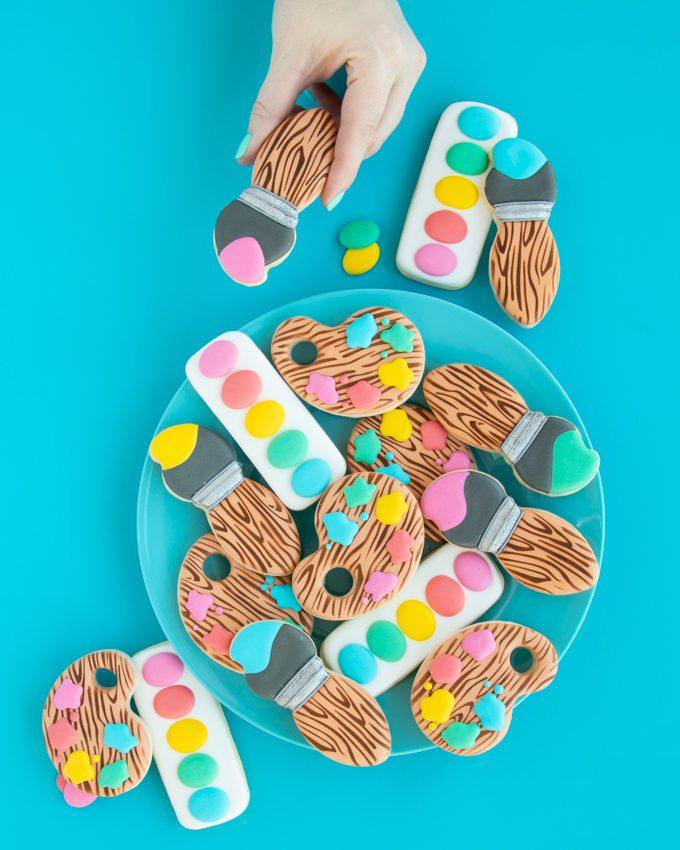 Various colors of edible food paint and brushes laid out next to plain butter cookies, ready for artistic decoration
