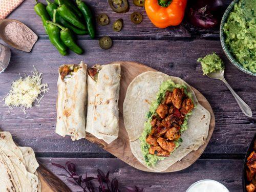 Overhead shot of two perfectly rolled chicken wraps on a rustic wooden board, ingredients scattered around