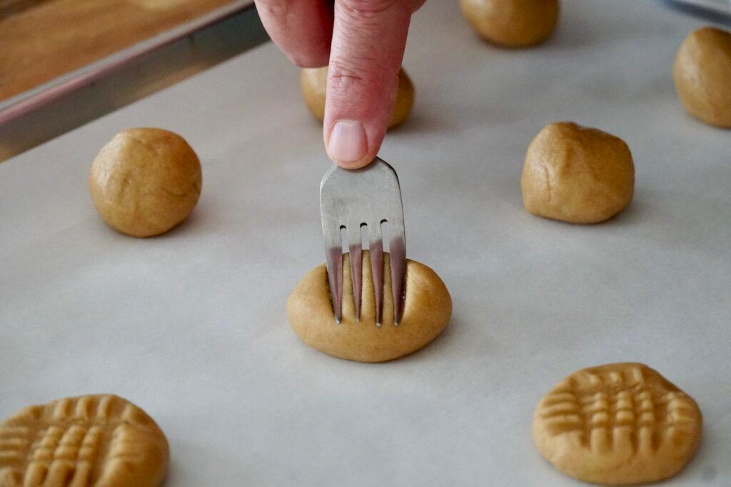 Close-up of hands pressing a fork into a peanut butter cookie dough ball on a baking sheet