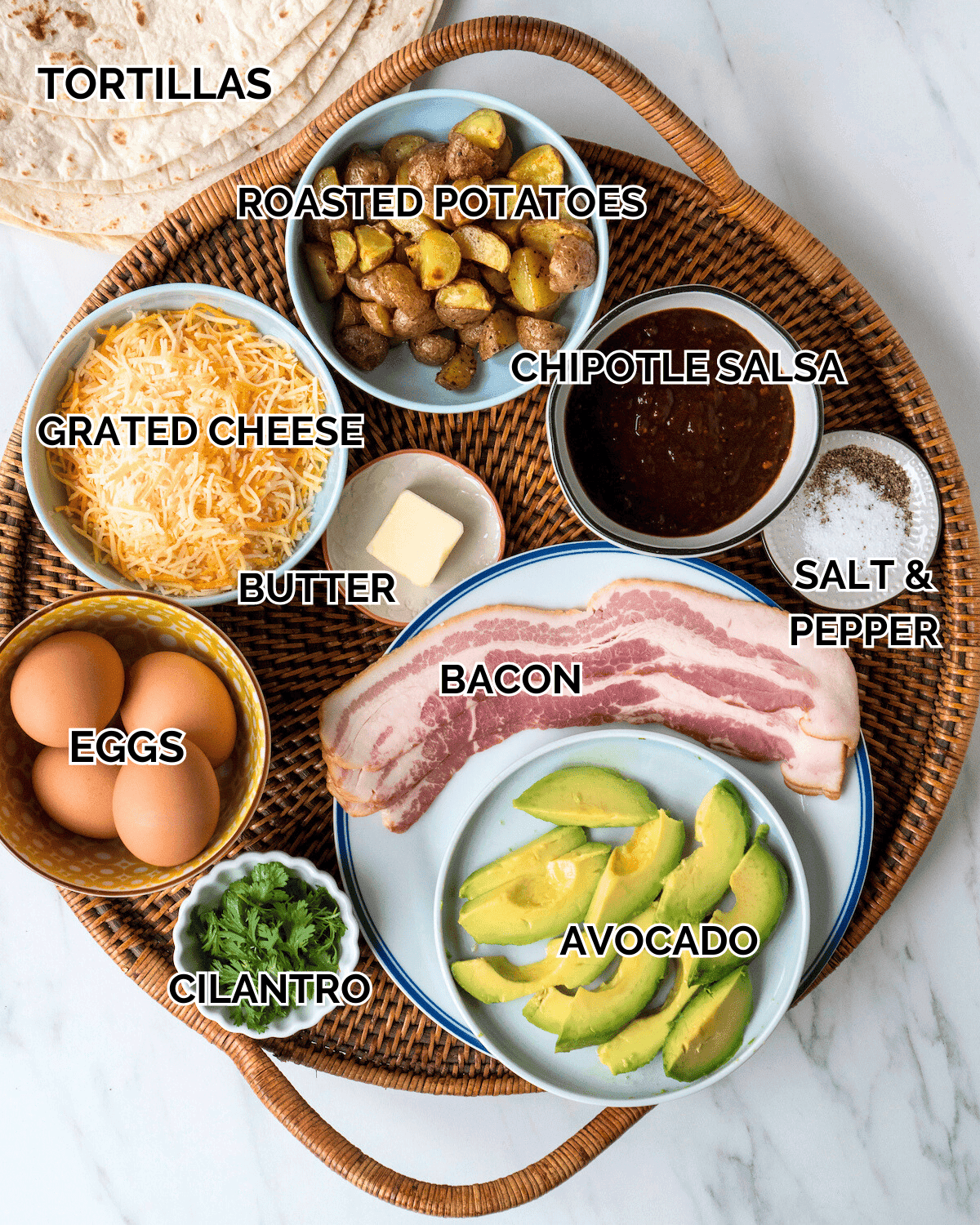 Ingredients for breakfast burritos laid out on a wooden cutting board: eggs, cheese, avocado, tortillas, salsa, red onion, cilantro, lime