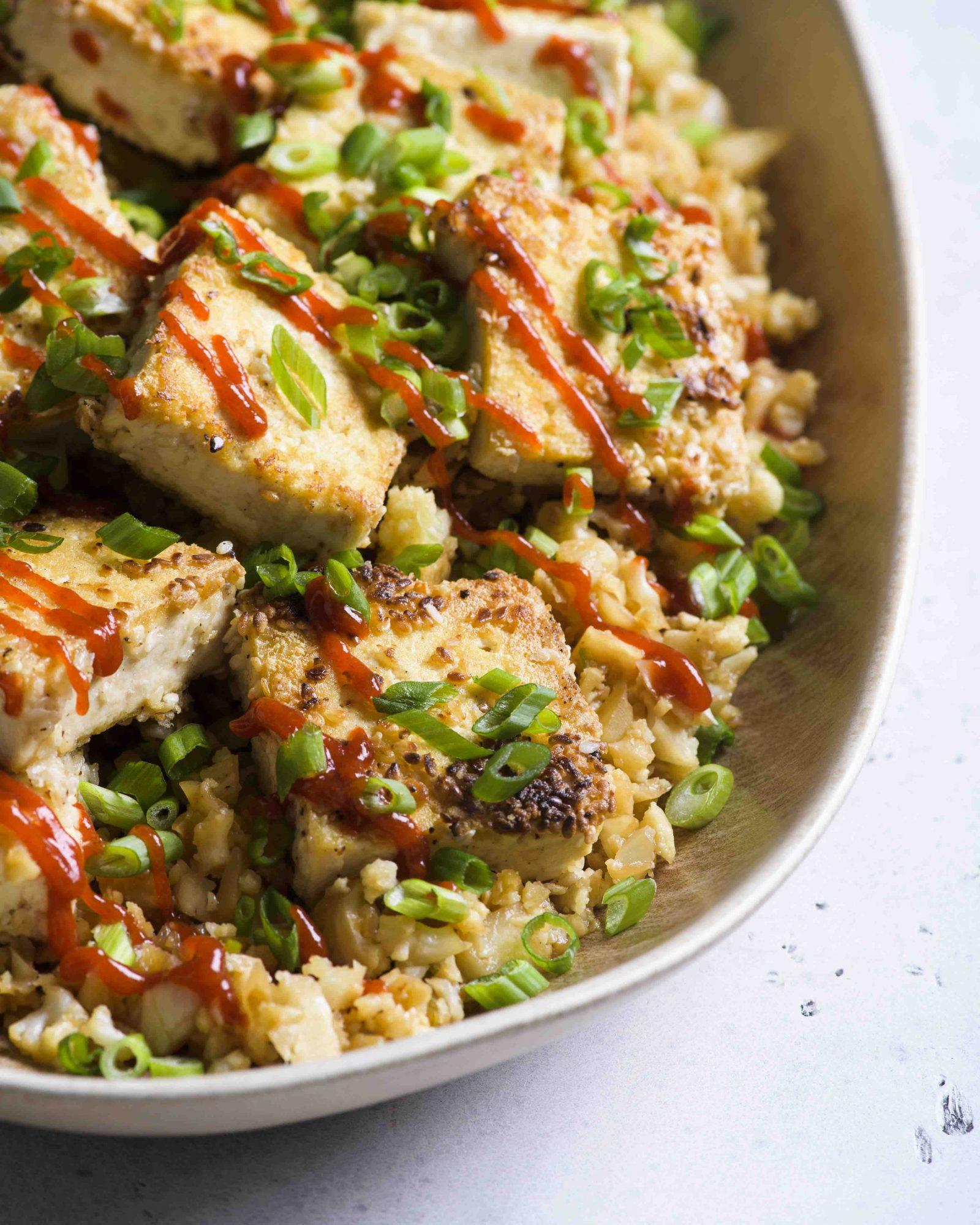 Close-up shot of a bowl of cauliflower rice stir-fry with tofu, garnished with sesame seeds and green onions
